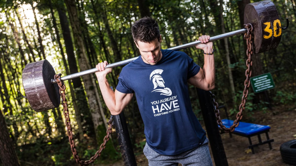 Man lifting barbell with chains in a wooded outdoor gym, showcasing strength training.